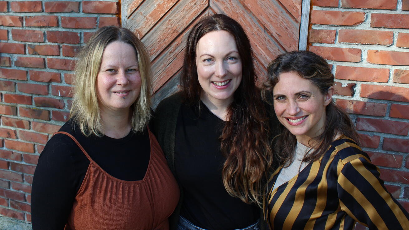 Tre damer framfor murvegg